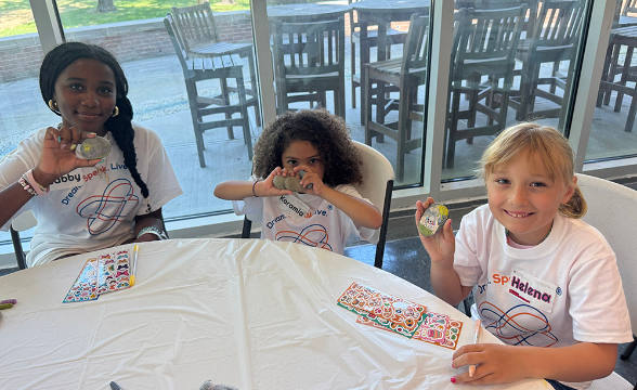 Three diverse girls in matching t-shirts smile while holding up decorated rocks at a craft table.