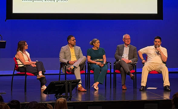 Five people sit on stage for a panel discussion, with one man holding a microphone. A large screen is behind them.