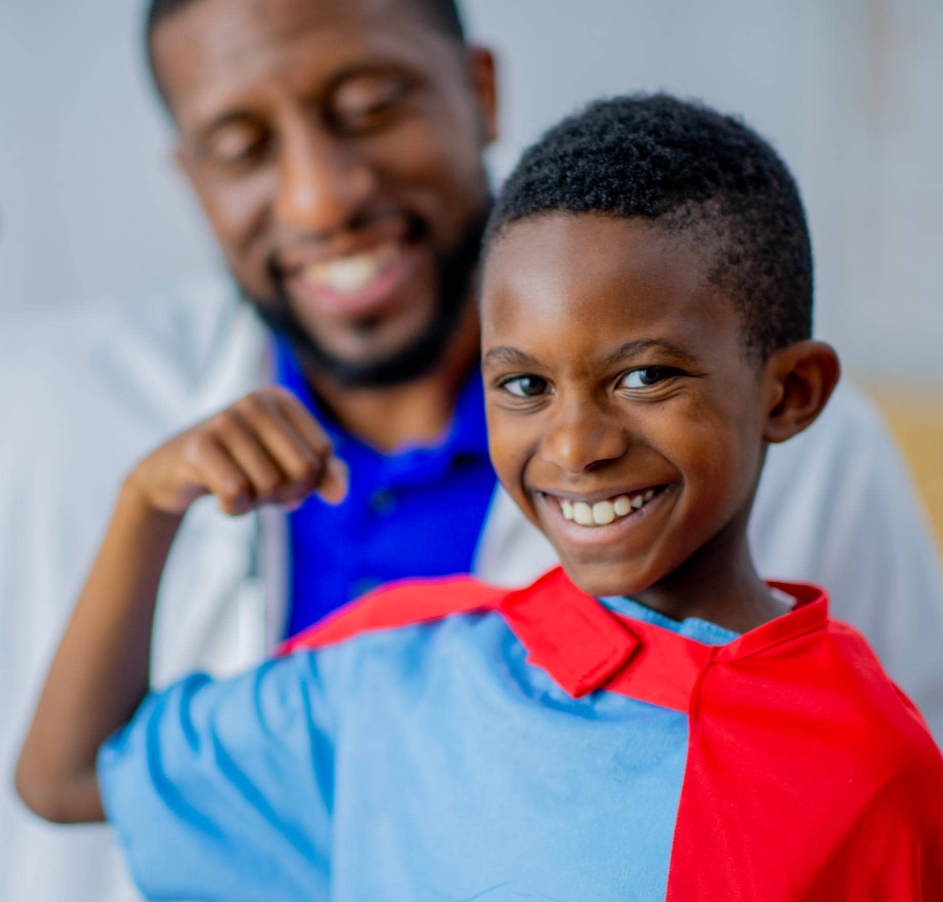 A smiling young boy wearing a blue shirt and red cape poses in front of a smiling male doctor in a white coat who is playfully mimicking a superhero pose.