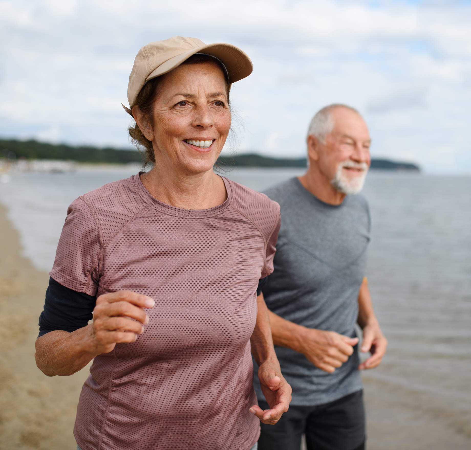 Two older adults jogging on a beach, with the woman in front wearing a cap and athletic shirt, smiling as they run along the shoreline.