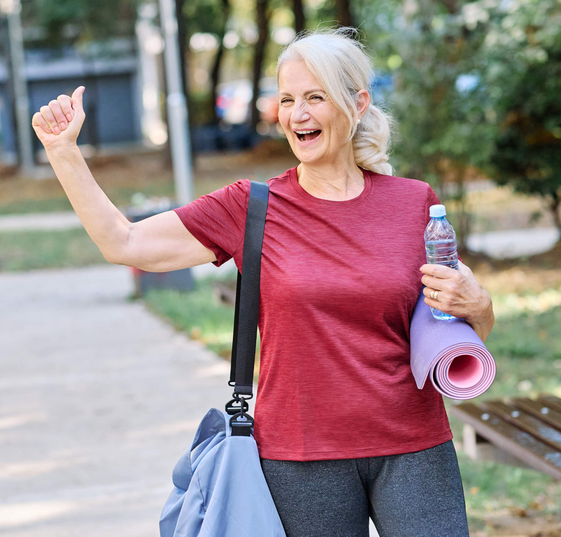 An older woman with grey hair in a ponytail, wearing a red shirt and holding a yoga mat and water bottle, smiles and gives a thumbs up outdoors.
