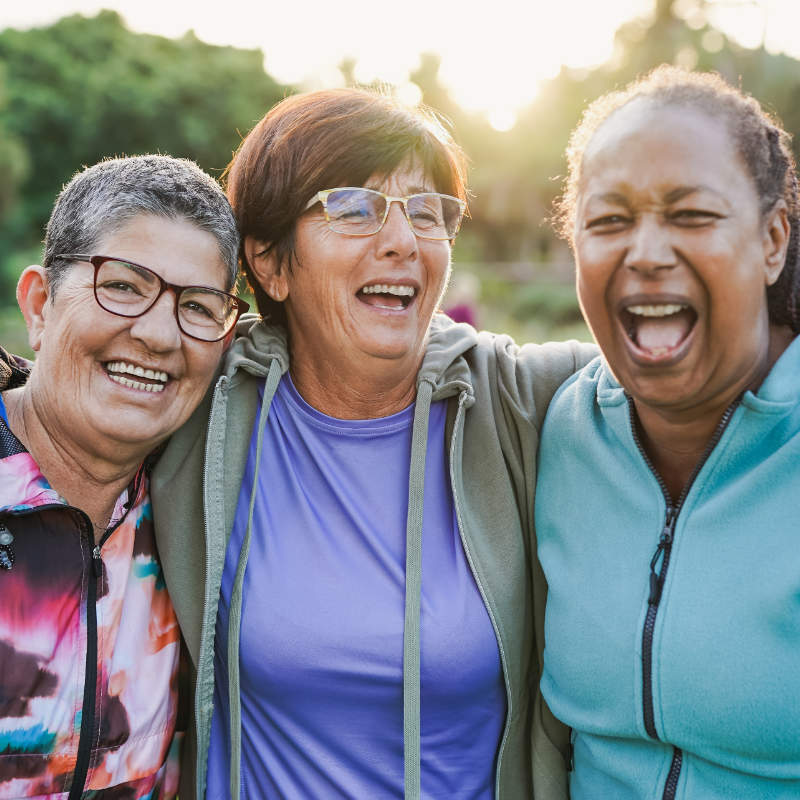 Three women laughing together outdoors, arms around each other, with sunlight in the background.