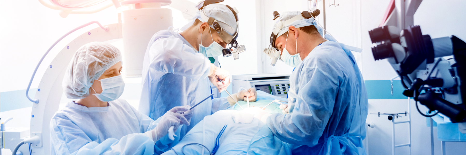 Three surgeons in blue scrubs, masks, and headlamps performing surgery in a modern operating room, with medical equipment visible in the background.