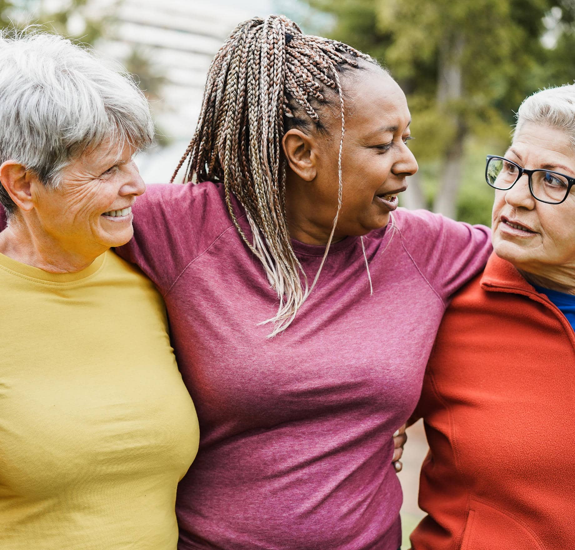 Three diverse older women standing close together outdoors, smiling and laughing. The woman in the center has braided hair and wears a maroon shirt, with her arms around her two friends—one in a yellow top and the other in an orange jacket with glasses.