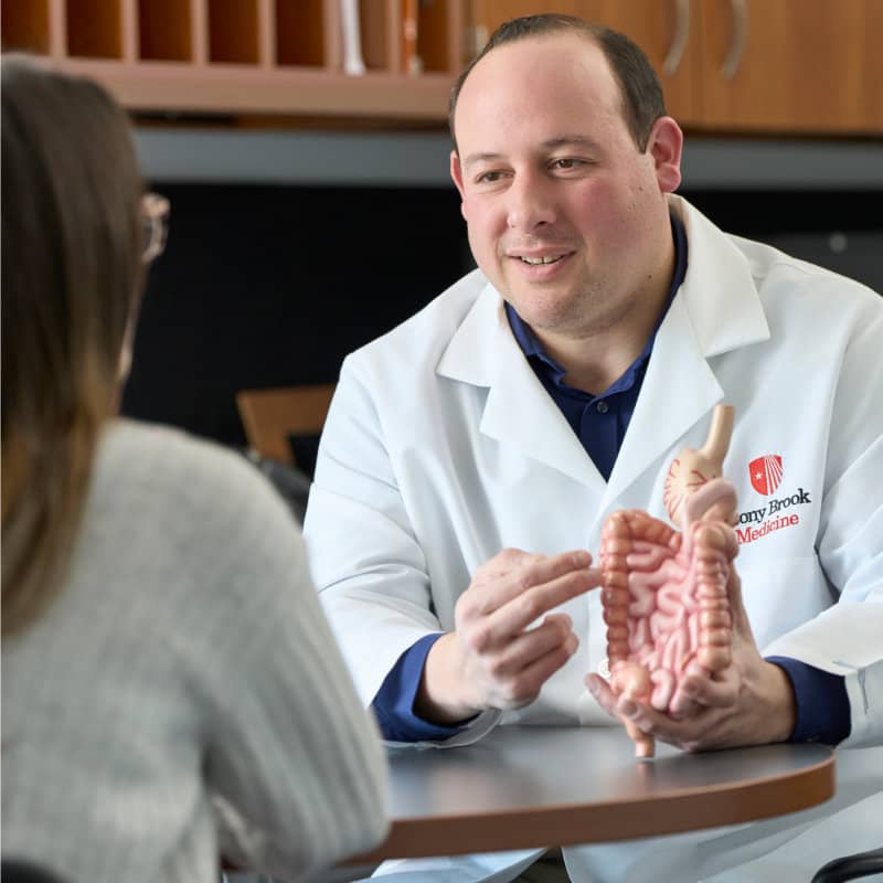 A male doctor in a white lab coat with a "Stony Brook Medicine" logo explains a model of the human digestive system to a patient during a consultation.