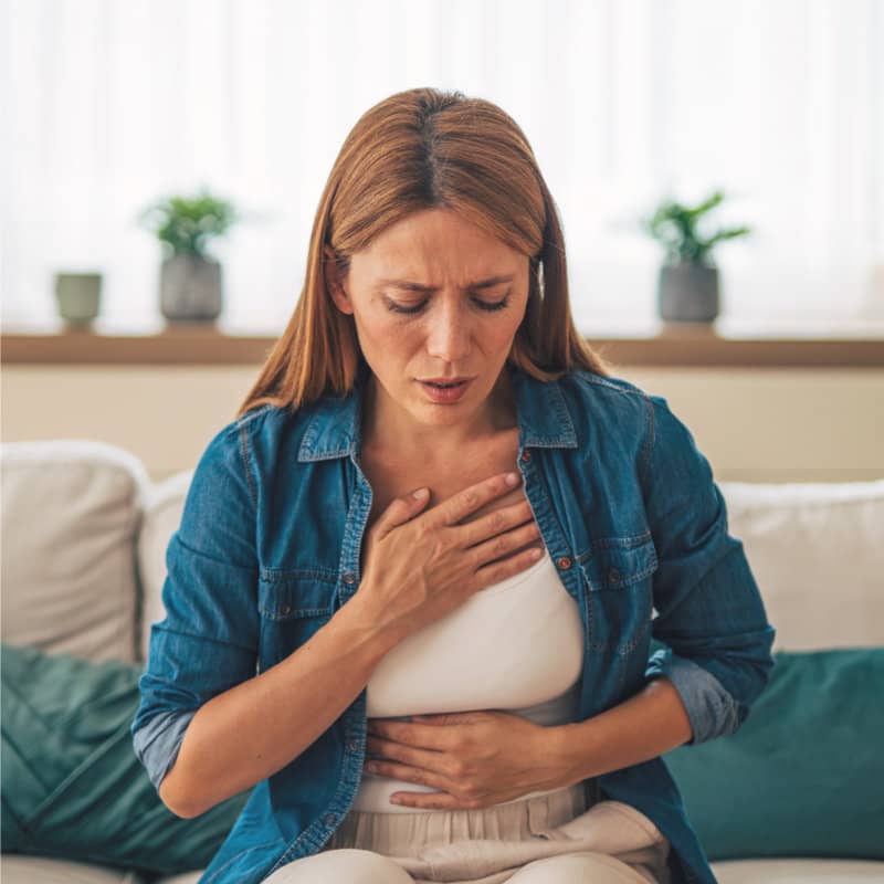 A woman with reddish-brown hair sits on a couch, looking distressed and holding her chest and stomach, indicating discomfort or pain.