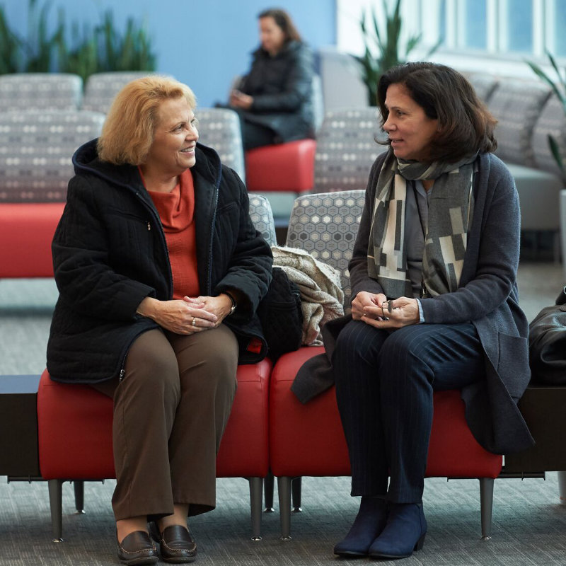 Two women talking while sitting on red chairs in a waiting area.