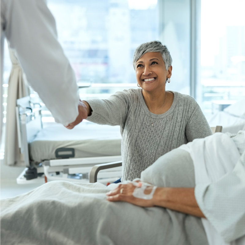 A smiling female patient in a hospital bed shakes hands with a doctor in a bright hospital room.