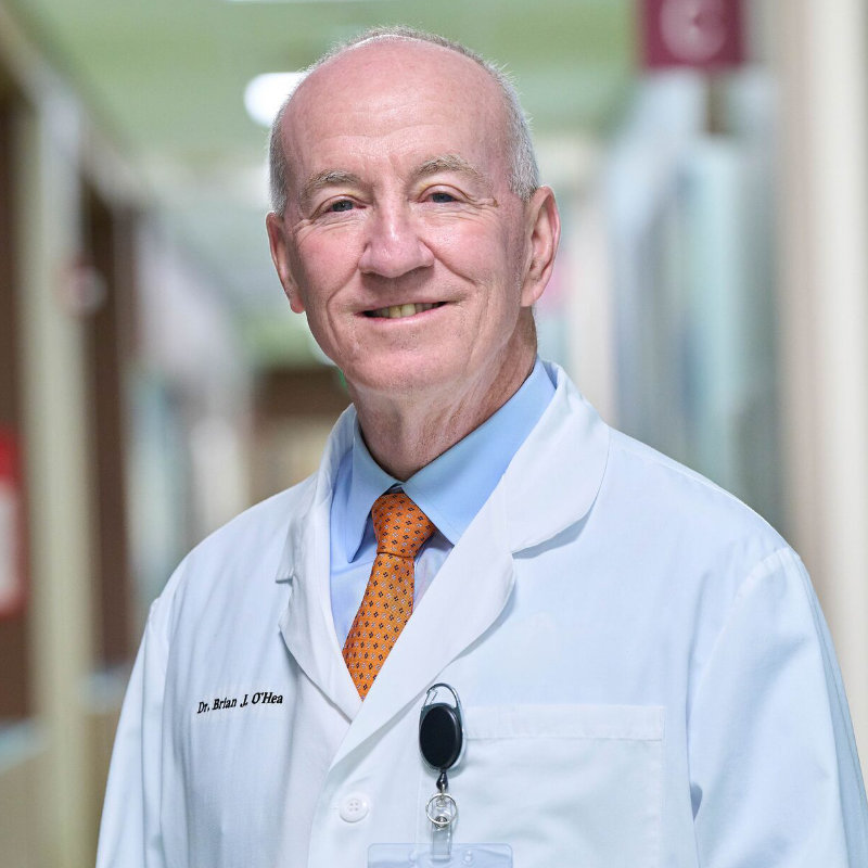 A male doctor in a white coat with an orange tie smiles in a hospital hallway.