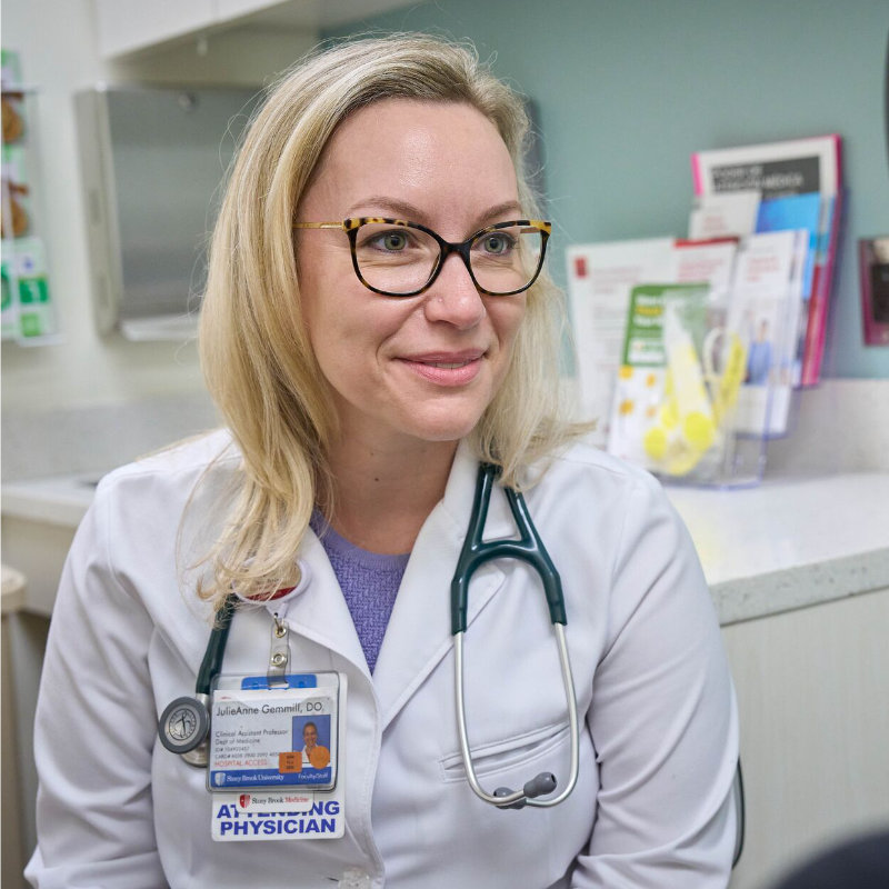 A female doctor with blonde hair and glasses, wearing a white coat and stethoscope, smiles in an office.