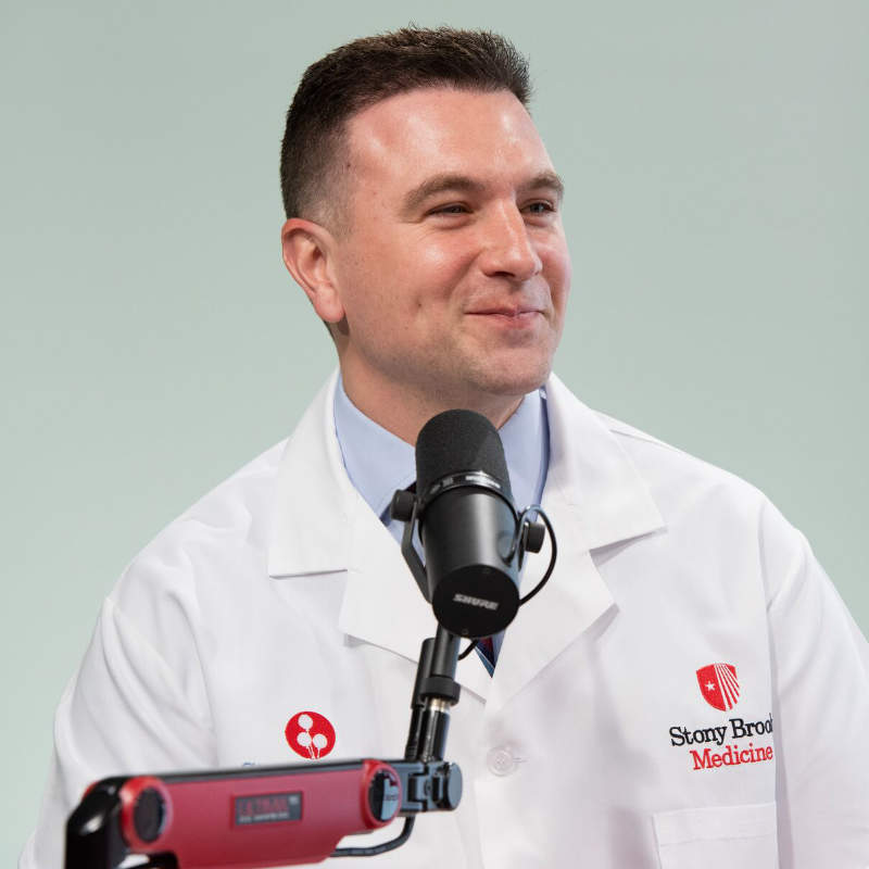 A male doctor in a white lab coat with a "Stony Brook Medicine" logo speaks into a microphone.