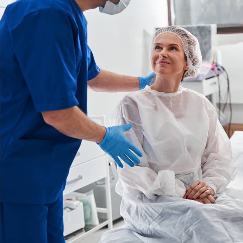 A female patient in hospital gown and hair cover sits on medical bed while a healthcare worker in blue scrubs stands beside her.
