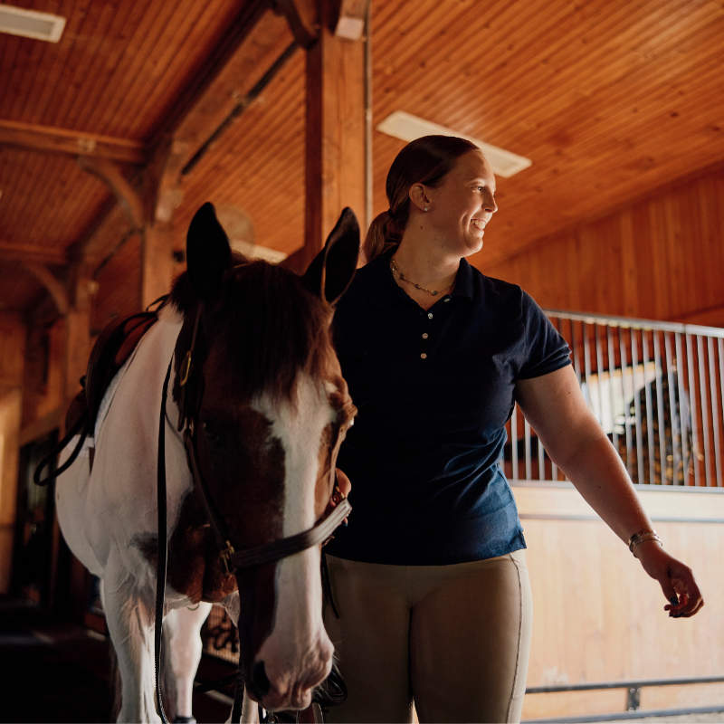 A smiling woman with her hair pulled back stands inside a wooden stable next to a brown and white horse. The horse is wearing a saddle and bridle, and the woman is looking off to the side.