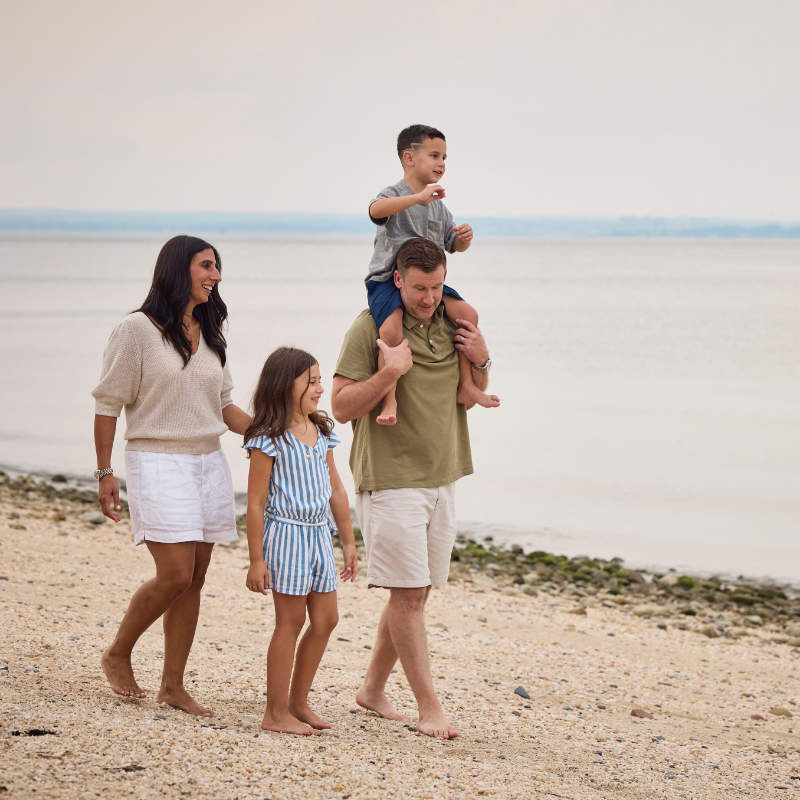 A family of four walking along a pebbled beach. The father is carrying his young son on his shoulders, while the mother walks beside him holding hands with their young daughter. They are all smiling and looking towards the water.