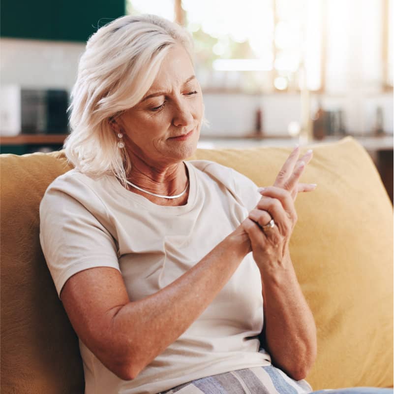 An older woman with white hair sits on a couch, holding her wrist as if in pain.