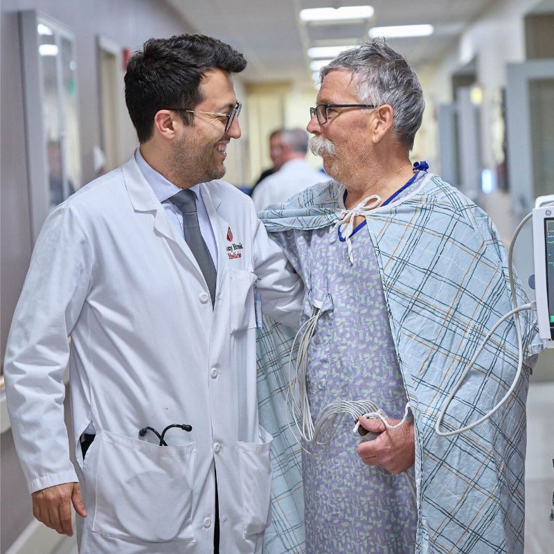 A doctor in a white coat chats warmly with a male patient in a hospital gown in a hospital corridor.