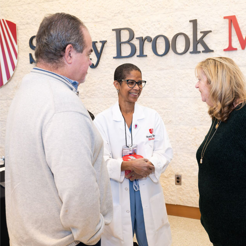 A Stony Brook Medicine staff member in a lab coat speaks with two visitors in a hospital lobby.