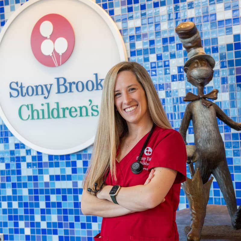 A smiling female healthcare professional in red scrubs stands with arms crossed in front of a blue tiled wall, next to a "Stony Brook Children's" sign and a bronze statue of the Cat in the Hat.