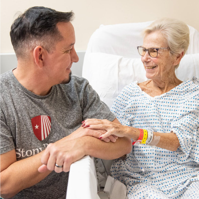 A man holds hands with an elderly woman in a hospital bed, both smiling at each other.