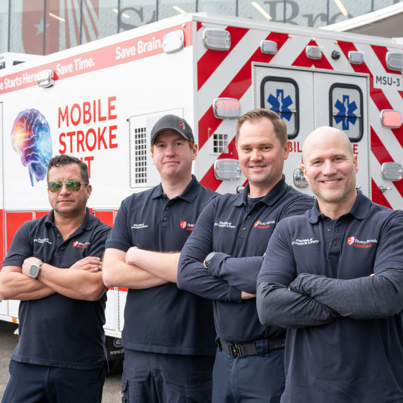 Four men in matching navy blue polo shirts standing with crossed arms in front of a 'Mobile Stroke Unit' ambulance, featuring red and white striping and medical insignia.