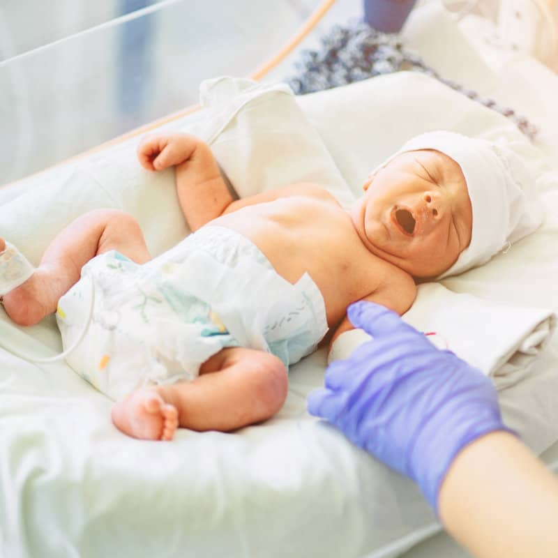 A newborn baby wearing a diaper and white cap lies in a hospital incubator, yawning widely while a gloved hand gently touches its arm.