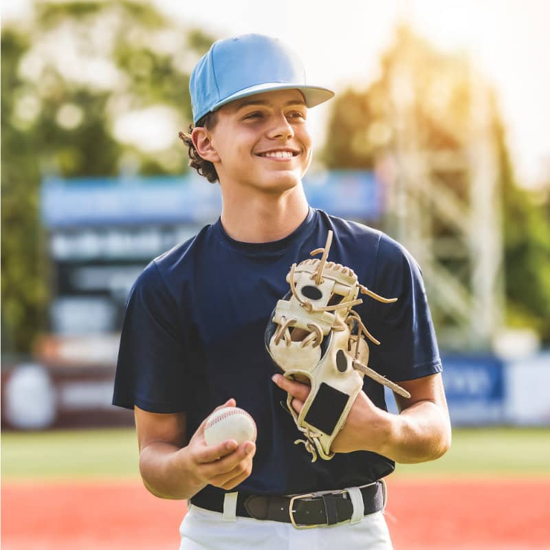 A smiling teenage boy in a blue baseball uniform and cap holds a baseball and glove on an outdoor field during golden hour.