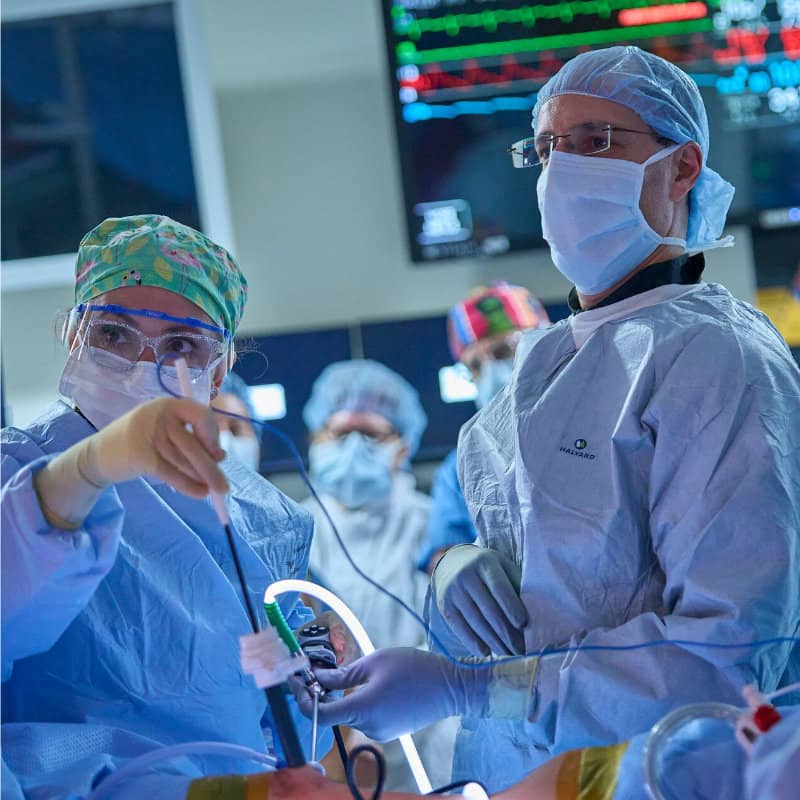 Two surgeons in full surgical gear, including masks and caps, work together in an operating room with medical monitors visible in the background.