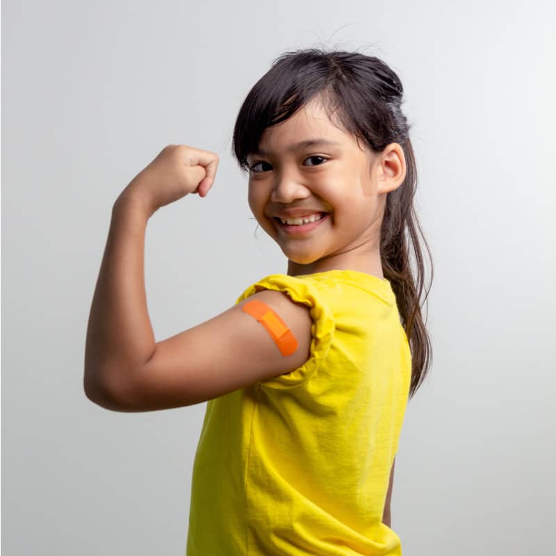 A smiling young girl in a yellow shirt flexes her arm to show an orange bandage on her upper arm against a plain background.