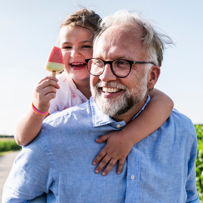 A grandfather carries his smiling granddaughter on his shoulders as she holds up a watermelon popsicle.