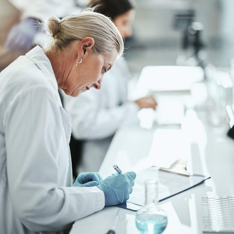 An older female scientist in a lab coat and blue gloves writes notes on a clipboard in a laboratory.