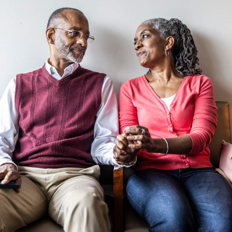 An older couple sits side by side, holding hands and smiling at each other in a warm, intimate moment.