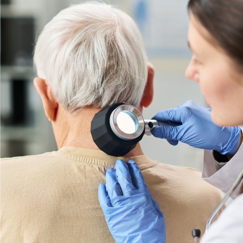A healthcare professional in blue gloves uses a large magnifying lamp to examine a patient's neck.