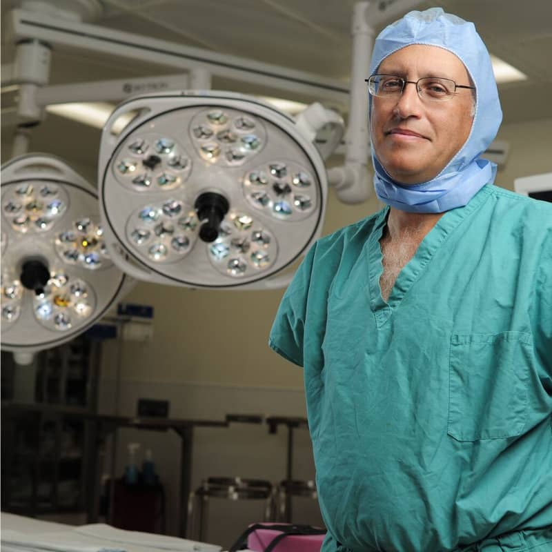 A male surgeon in green scrubs and surgical cap stands in an operating room beneath overhead surgical lights.