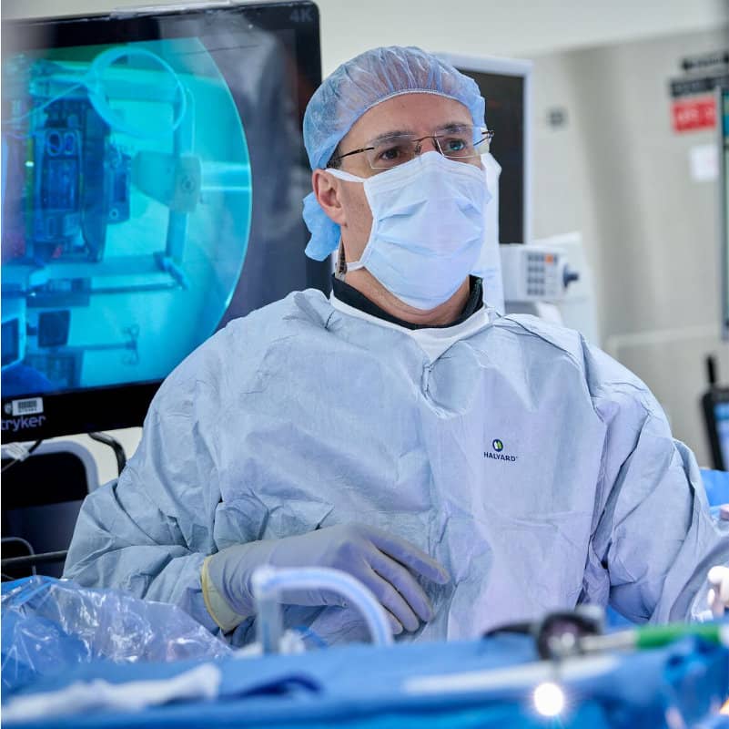 A surgeon in full surgical gear looks towards a monitor displaying an X-ray image.