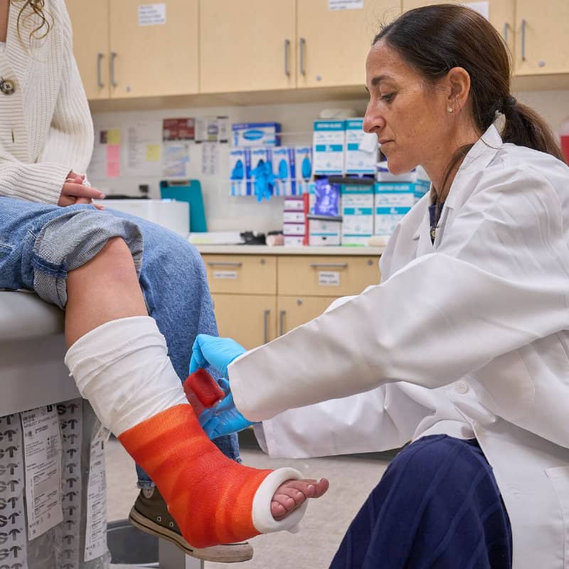 A medical professional in a lab coat carefully wraps a bright orange cast around a patient's leg.