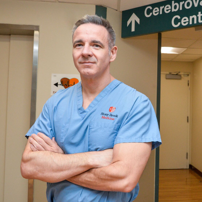 Dr. Fiorella in blue scrubs with crossed arms stands in a hospital corridor near a Cerebrovascular Center sign.