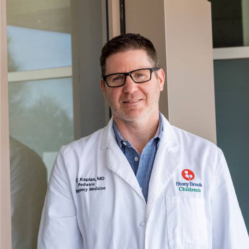 A male doctor wearing glasses and a white lab coat with "Stony Brook Children's" embroidery stands in front of a window, smiling at the camera.