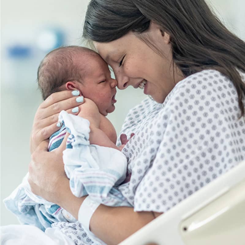 A smiling mother in a hospital gown holds her newborn baby, touching foreheads in a tender moment.