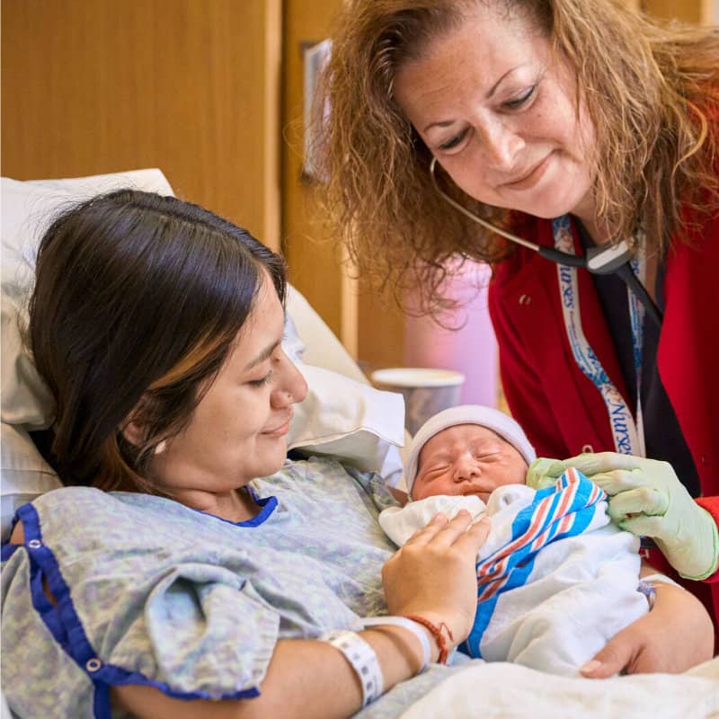 A nurse in red scrubs and gloves gently examines a newborn baby while the mother looks on from a hospital bed.