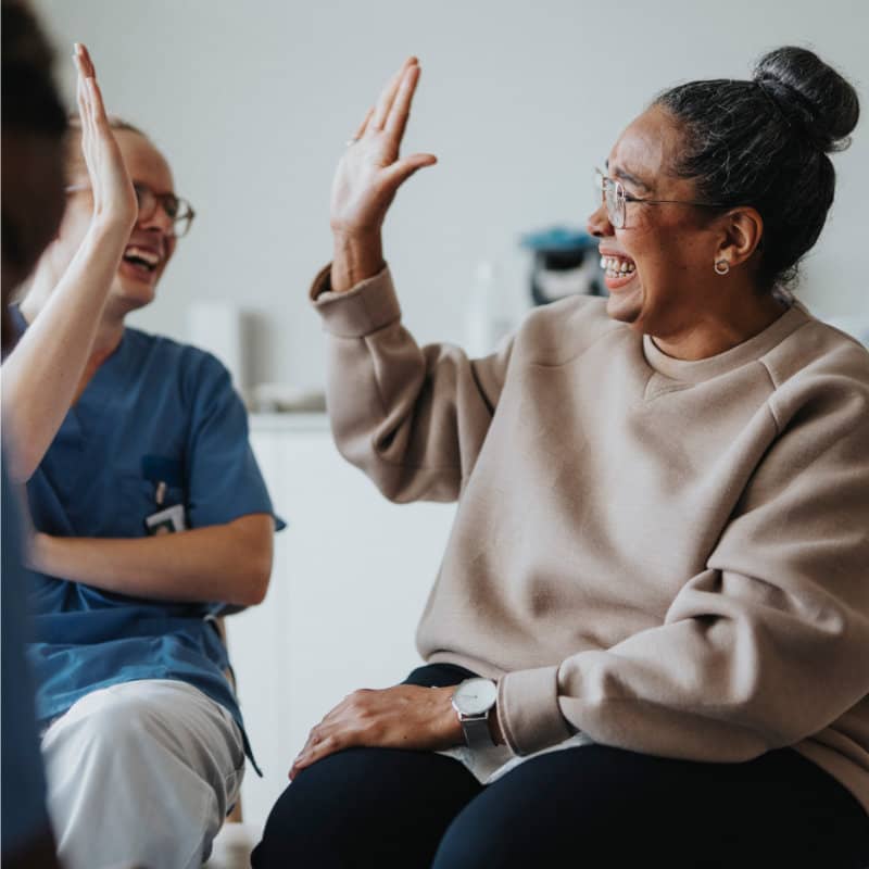 A smiling woman with glasses and her hair in a bun gives a high-five to a healthcare worker in blue scrubs during a therapy session.