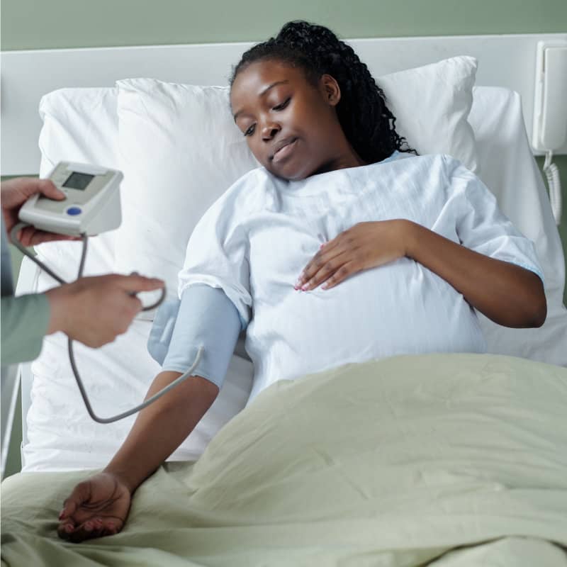 A pregnant woman lies in a hospital bed while a healthcare provider checks her blood pressure with a digital monitor.