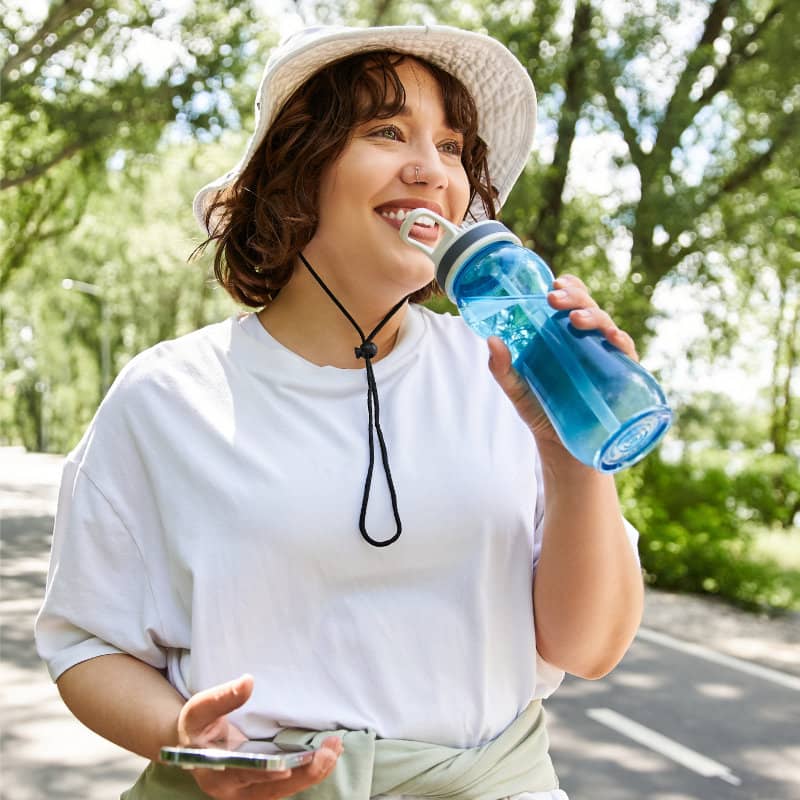 A woman in a white hat and t-shirt drinks from a blue water bottle while standing outdoors in a park.