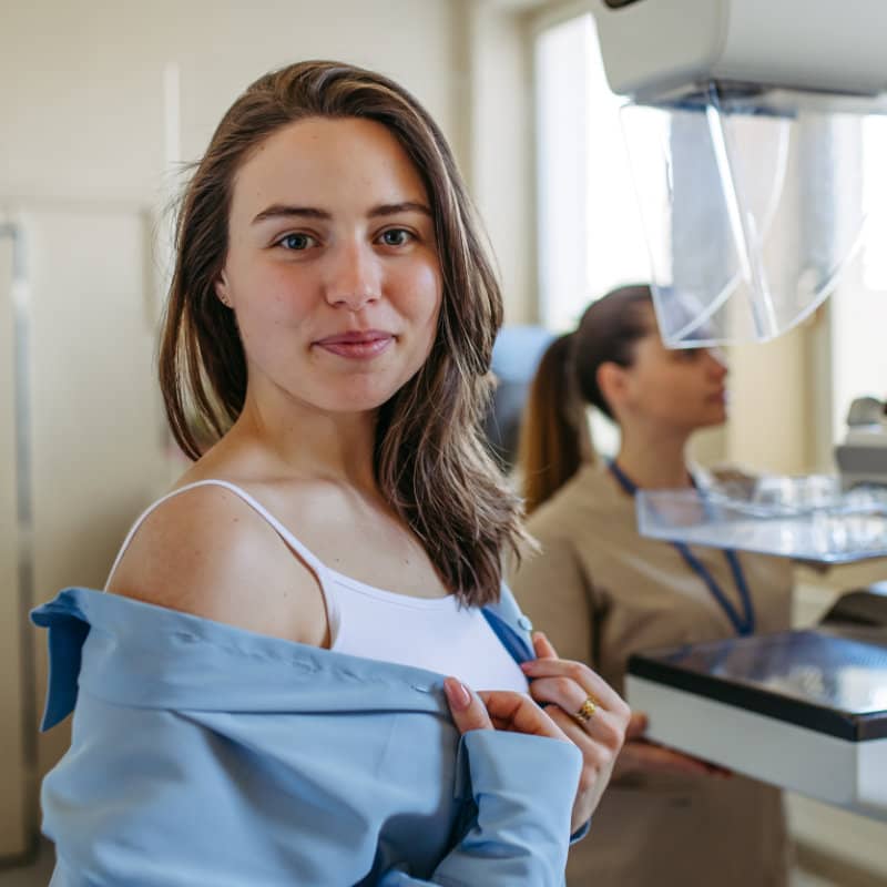 A young woman in a medical imaging room, holding open a blue gown while looking at the camera. A mammography machine and healthcare professional are visible in the background.