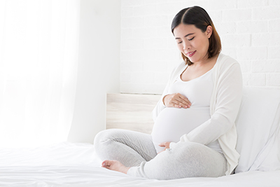 mujer asiática embarazada sosteniendo su vientre mientras cruza las piernas y se sienta