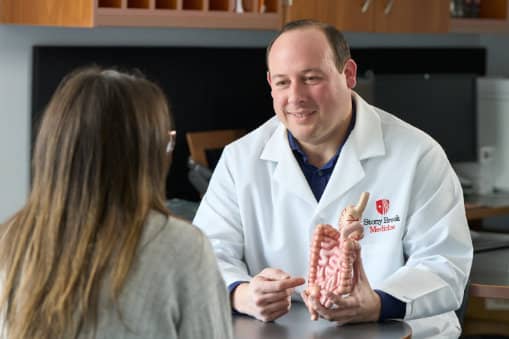 A doctor in a white lab coat from Stony Brook Medicine consulting with a patient while holding an anatomical model of the large intestine.