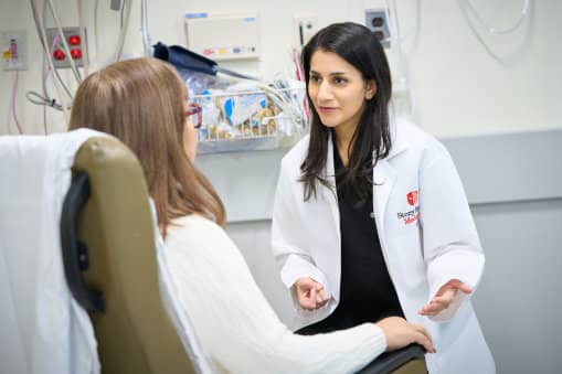 A female doctor in a white Stony Brook Medicine lab coat speaking with a seated patient in an examination room.