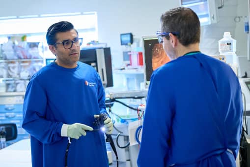 Two medical professionals in blue surgical gowns and gloves discussing a procedure in a clinical setting, with one holding an endoscope instrument.