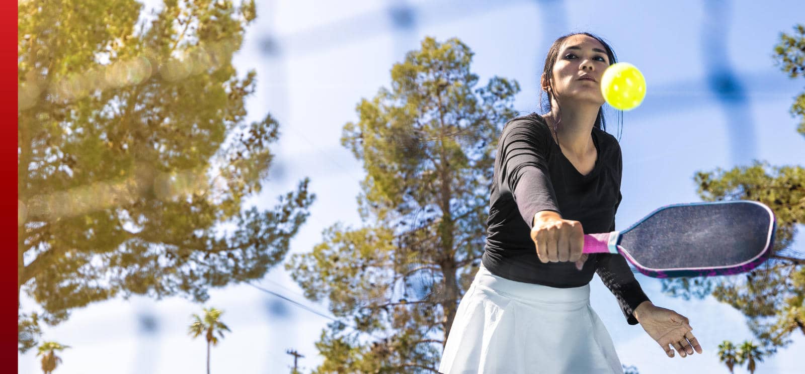 A woman in a black shirt and white skirt swings a pickleball paddle to hit a yellow ball on an outdoor court under a blue sky.