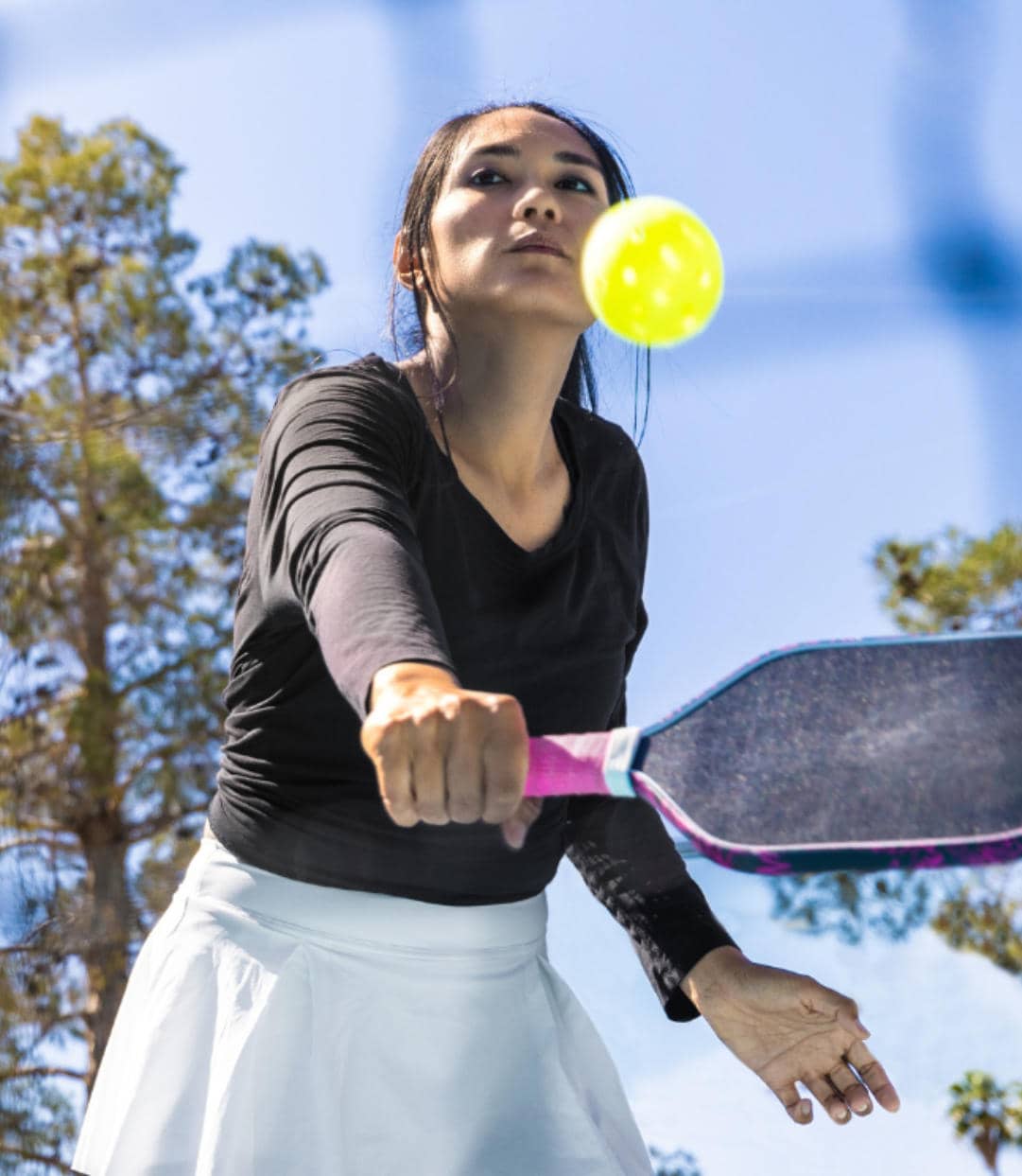 A woman in a black shirt and white skirt swings a pickleball paddle to hit a yellow ball on an outdoor court under a blue sky.